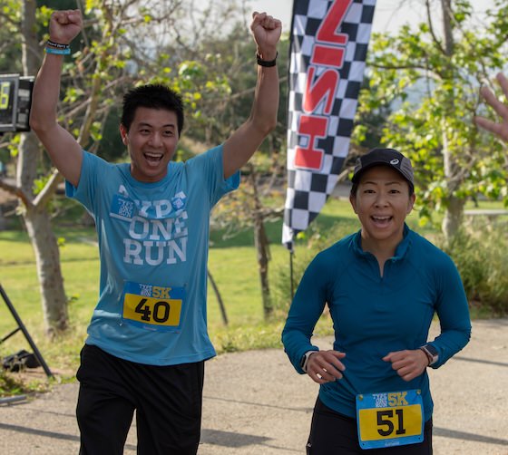 Two runners crossing the finish line at a 5K race, the man raising both fists in triumph while both smile with excitement