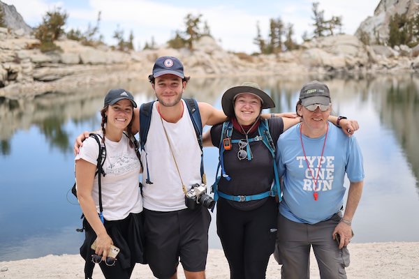 Four hikers with backpacks posing arm in arm at an alpine lake surrounded by granite rocks and scattered pine trees